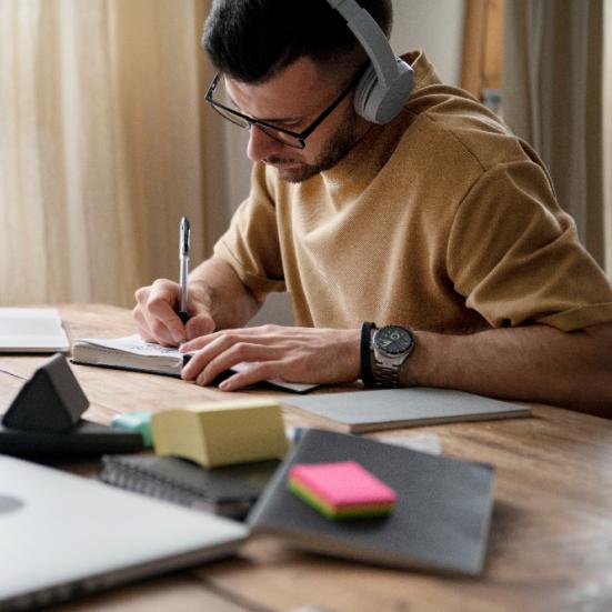 Mand, der skriver på en notesblog. Der ligger papirer, post-its og en computer rundt på skrivebordet.