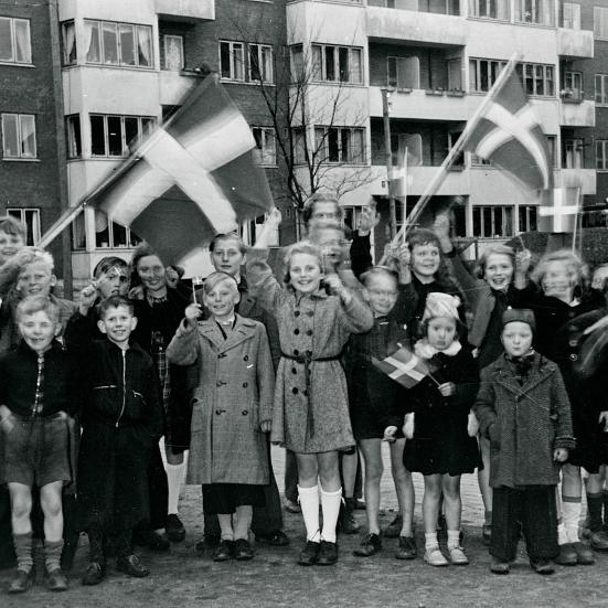Foto i sort/hvid af en masse børn, der smiler og vifter med dannebrogsflag foran en lejlighedsbygning.
