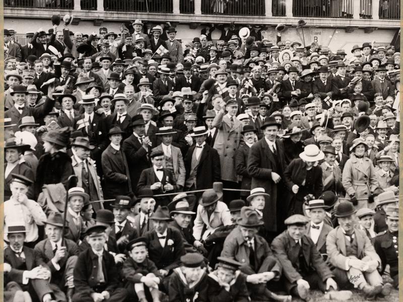Fotografi i sort / hvid af en stor gruppe mennesker. De er alle nydeligt klædt på, og bærer hatte. De er til fodboldkamp udenfor, og står i tilskuerrækker.