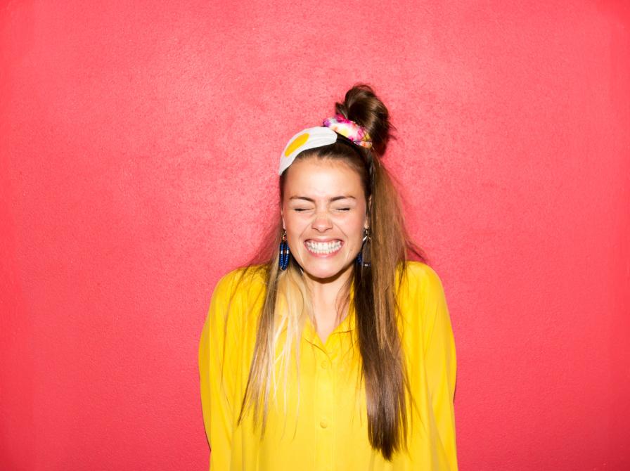 Kvinde med langt hår og gult tøj smiler med lukkede øjne.