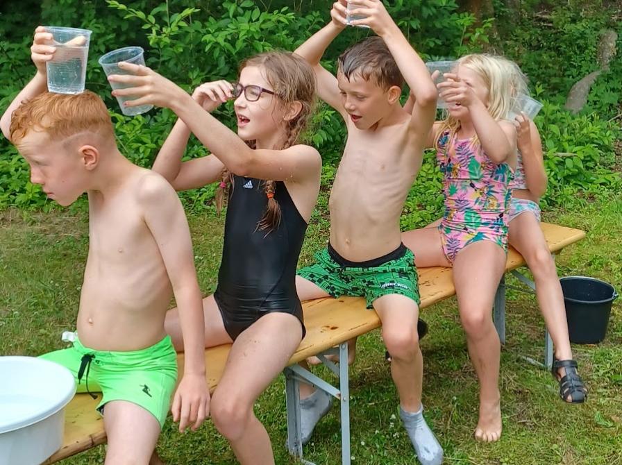 Fem børn sidder på en bænk i badetøj. De leger med vand i vandglas. 